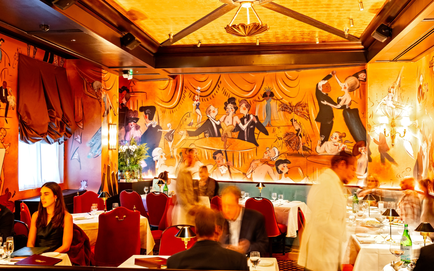 The opulent dining room at dinner service, with red velvet chairs, white tablecloths, and a sweeping art deco mural