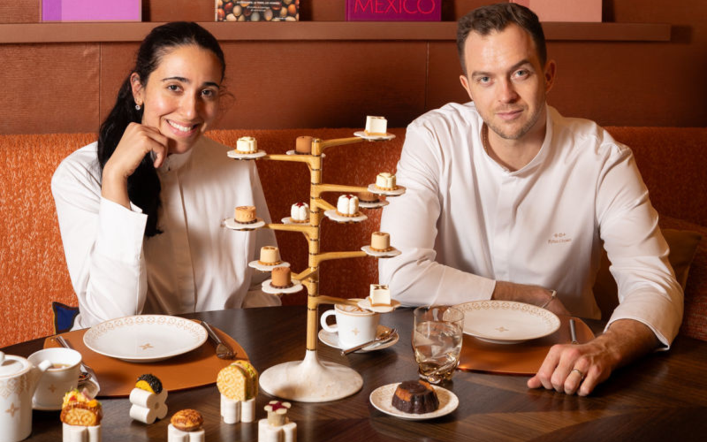 Two chefs seated alongside an elaborate tiered petit four display and sculptural tea service at Le Cafe