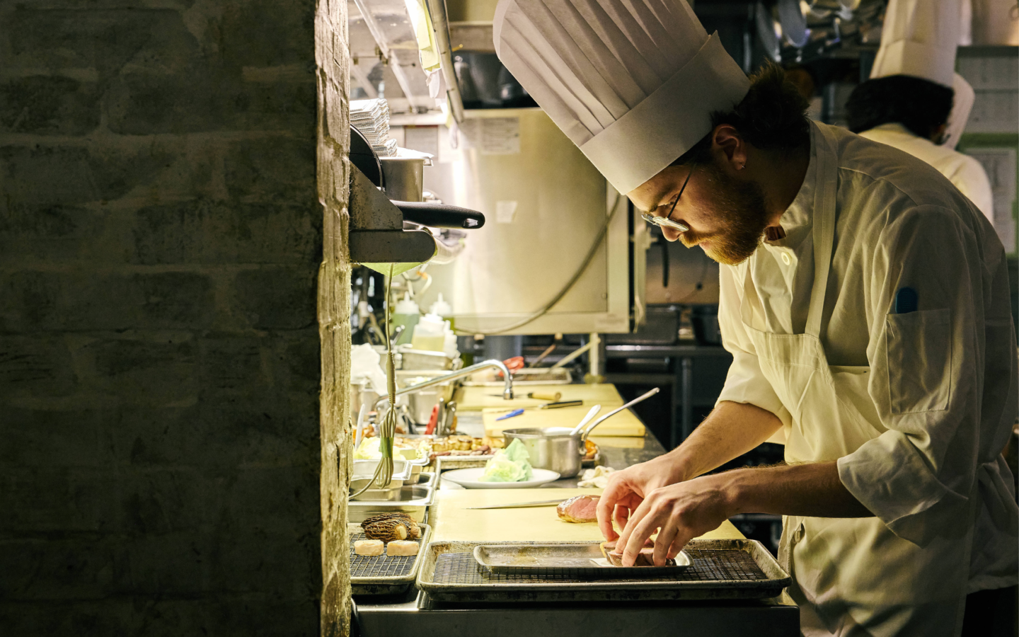Le Coucou chef in a white toque carefully plating a dish during dinner service in a warmly lit kitchen